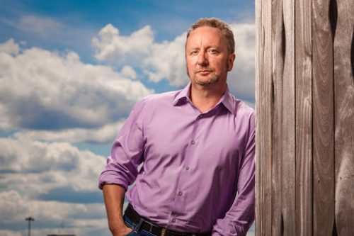 Caucasian man in purple shirt leaning against wood wall outdoors on blue-skyed day with clouds.