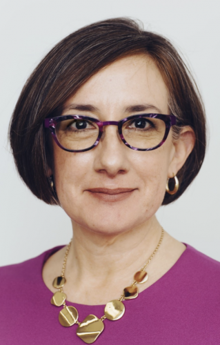 White woman with glasses, short brown hair, ornate necklace, magenta shirt, and round earrings.