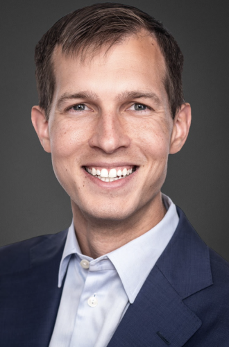 White man, smiling, baby blue button-down shirt, navy blue blazer, grey background