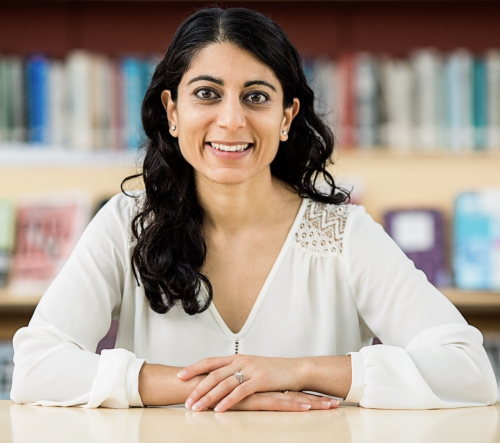 Woman with dark curly hair, white shirt, smiling, books in the background
