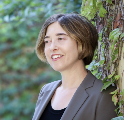 White woman smiling. She is wearing grey blazer and a black shirt. Tree in the background