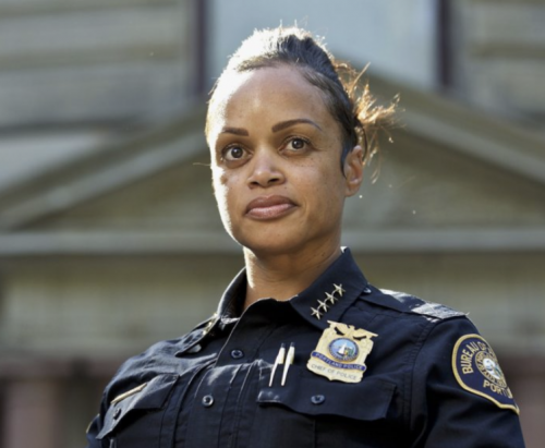 Black woman in a navy blue police uniform. Background is a blurry building.
